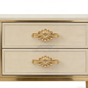 Puede incluir: Primer plano de un mueble de dos cajones con acabado crema y ribetes dorados. Cada cajón presenta un tirador dorado en forma de flor. El diseño del mueble es clásico y elegante, adecuado para la decoración del dormitorio o salón.