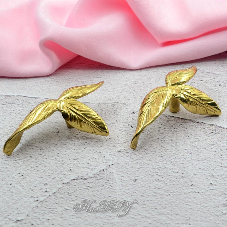 Pu&ograve; includere: Due elementi decorativi dorati a forma di foglia. Ogni pezzo ha tre foglie con venature dettagliate. L'hardware &egrave; posto su uno sfondo rosa tenue.