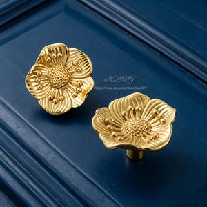 May include: Two gold-colored, flower-shaped cabinet knobs. Each knob features a detailed floral design with a textured center and petal detailing. The knobs are set against a dark blue background. The text "HaoDIY" and a website address are visible.