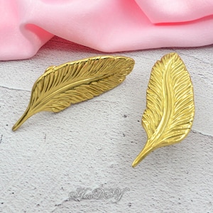 Peut inclure: Deux objets d&eacute;coratifs dor&eacute;s en forme de plumes. Les plumes pr&eacute;sentent des veines d&eacute;taill&eacute;es et une forme l&eacute;g&egrave;rement incurv&eacute;e. Le fond est un tissu rose clair et une surface gris clair.