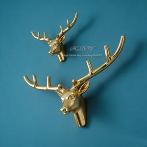 Puede incluir: Dos ganchos de pared con forma de cabeza de ciervo doradas. Los ganchos están montados en una pared azul turquesa. Las cabezas de ciervo tienen astas detalladas y miran en diferentes direcciones. El texto "HaoDIY" y una dirección web son visibles.