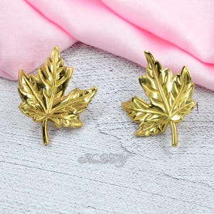 May include: Two gold-colored maple leaf-shaped decorative objects. The leaves have detailed veining and are set against a pink fabric and a white wooden surface. The objects appear to be made of metal.
