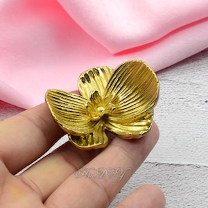 May include: A gold-coloured orchid flower brooch. The brooch is made of metal and has detailed petal and leaf textures. The brooch is held in a hand against a pink background. The text "Hao DIY" is visible.