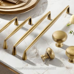 Op de afbeelding: Een verzameling antieke messing kastbeslagen, waaronder verschillende handgrepen en ronde knoppen. De handgrepen variëren in lengte en de knoppen hebben een afgerond ontwerp. De hardware wordt gepresenteerd op een wit marmeren oppervlak.