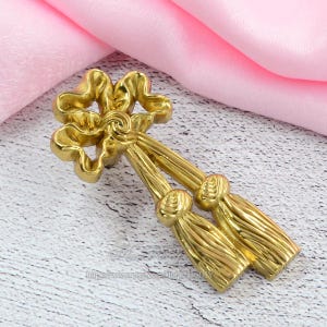 May include: A gold-coloured decorative tassel with a bow-like top. The tassel features two long, textured strands with decorative knots and fringe. The design is ornate and likely intended for interior decor or craft projects.