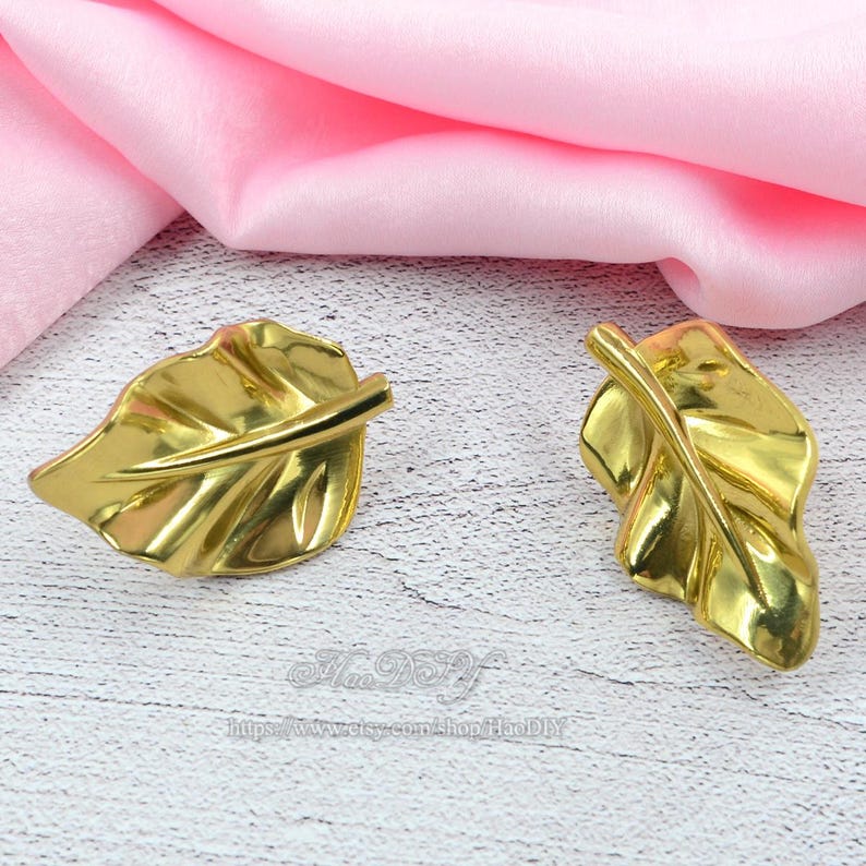 Op de afbeelding: Twee goudkleurige, bladvormige decoratieve objecten. De bladeren hebben gedetailleerde nerven en een glanzende afwerking. Ze worden gepresenteerd op een lichtgekleurd oppervlak met roze stof op de achtergrond. Het zijn waarschijnlijk decoratieve accessoires.
