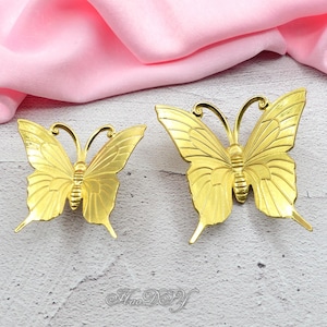 Pode incluir: Dois objetos decorativos dourados em forma de borboleta. As borboletas têm padrões de asas detalhados e antenas curvas. Os objetos são exibidos em uma superfície cinza claro com um tecido rosa ao fundo.