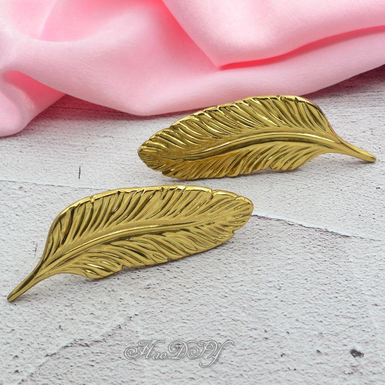 Peut inclure: Deux objets d&eacute;coratifs en forme de plumes dor&eacute;es. Les plumes pr&eacute;sentent des veines d&eacute;taill&eacute;es et un &eacute;clat m&eacute;tallique. Les objets sont probablement utilis&eacute;s pour la d&eacute;coration int&eacute;rieure ou les projets d'artisanat. Le fond est un tissu rose clair.