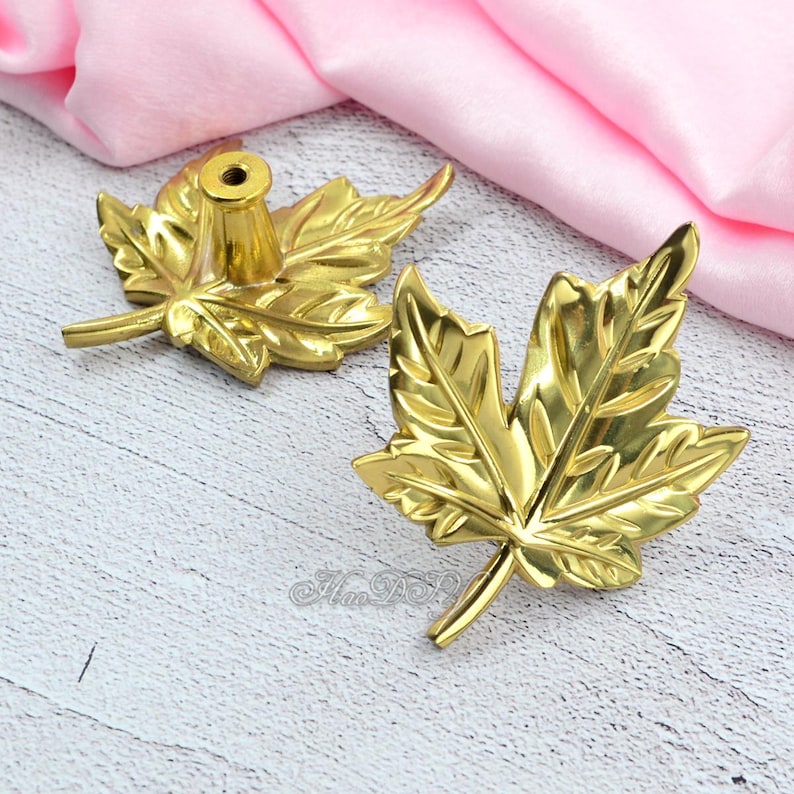 May include: Two gold-colored maple leaf cabinet knobs. One knob has a cylindrical base for mounting. The other knob is a decorative leaf. The knobs are on a light wood surface with a pink fabric background.