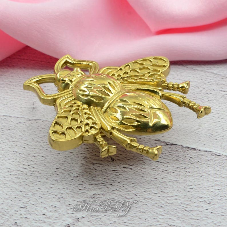 Peut inclure: Une broche dor&eacute;e en forme d'abeille. La broche pr&eacute;sente des ailes, des segments de corps et des pattes d&eacute;taill&eacute;s. Le design est tr&egrave;s d&eacute;taill&eacute; et semble &ecirc;tre en m&eacute;tal. La broche est pr&eacute;sent&eacute;e sur un fond en tissu rose.