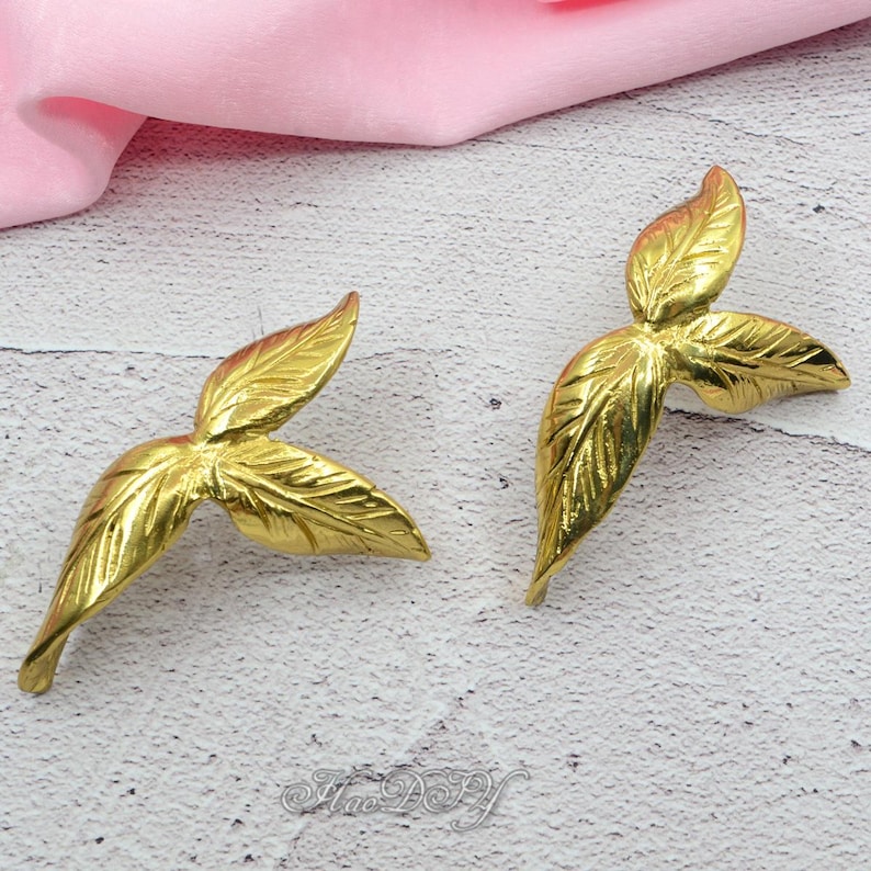 Pu&ograve; includere: Due abbellimenti dorati a forma di foglia. Ognuno ha tre foglie con venature dettagliate. Le foglie sono disposte a ventaglio. Lo sfondo &egrave; una superficie rosa chiaro e bianca.