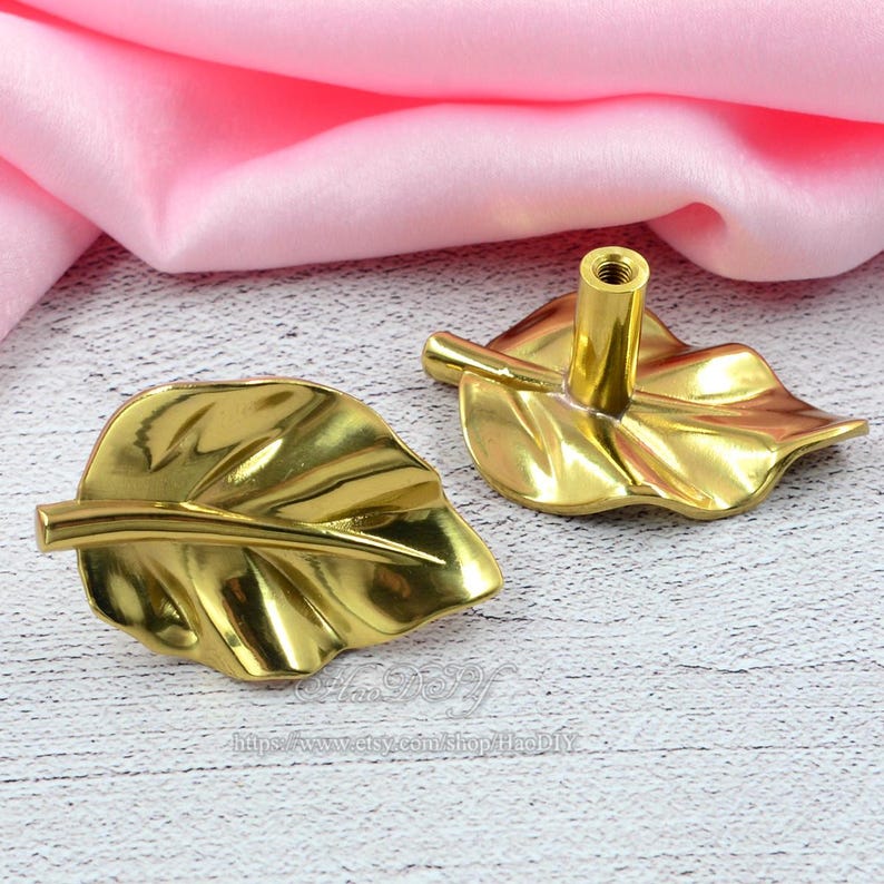 Op de afbeelding: Twee goudkleurige kastknoppen in de vorm van een blad. De knoppen hebben een gedetailleerd bladnervenontwerp en een steelachtige handgreep. De achtergrond is een lichtroze stof en een wit houten oppervlak.