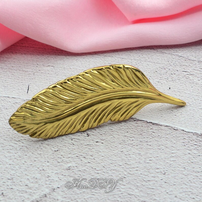Peut inclure: Une plume en m&eacute;tal dor&eacute; avec des veines d&eacute;taill&eacute;es. La plume est pos&eacute;e sur une surface blanche et grise avec un tissu rose en arri&egrave;re-plan. La plume est un objet d&eacute;coratif.