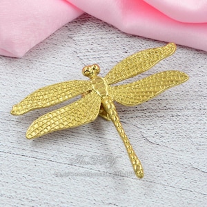 May include: A gold-coloured dragonfly brooch with textured wings and body. The dragonfly has detailed features, including eyes and segmented body. The brooch is lying on a light-coloured wooden surface with a pink fabric in the background.