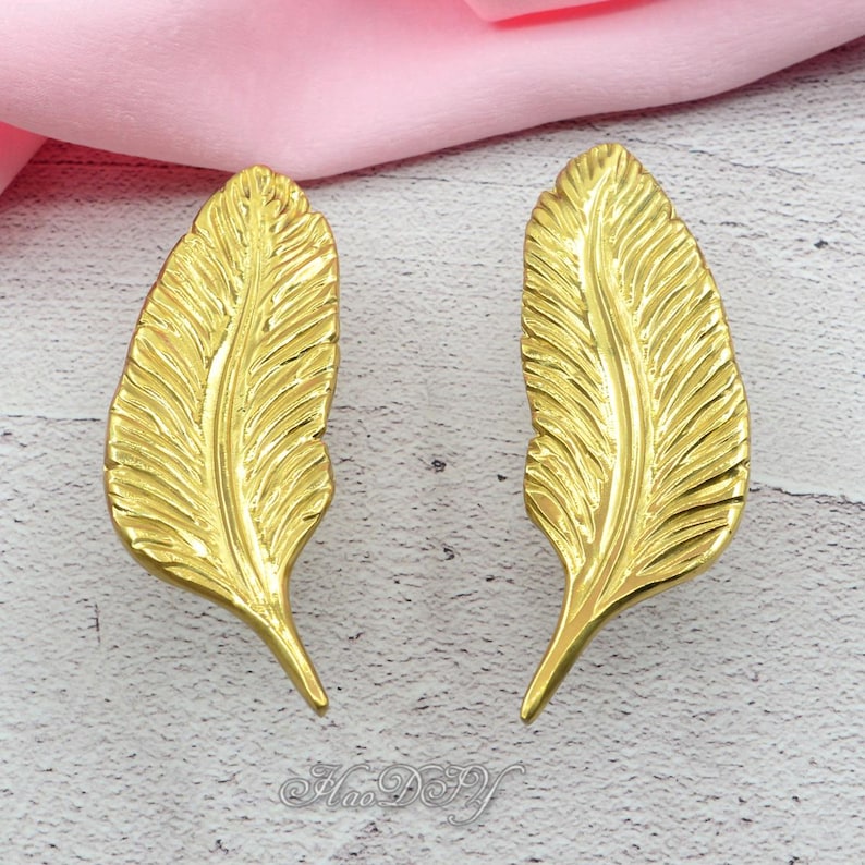 Peut inclure: Deux objets d&eacute;coratifs dor&eacute;s en forme de plumes. Chaque plume pr&eacute;sente des veines d&eacute;taill&eacute;es et une forme incurv&eacute;e. Les objets sont pr&eacute;sent&eacute;s sur une surface claire avec un tissu rose en arri&egrave;re-plan.