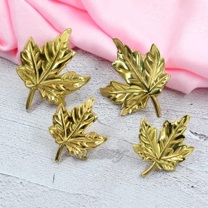 May include: Four gold-colored maple leaf-shaped decorative items. The leaves have detailed veining and a shiny finish. They are arranged on a light-colored wooden surface with a pink fabric in the background. The leaves are likely decorative accents.