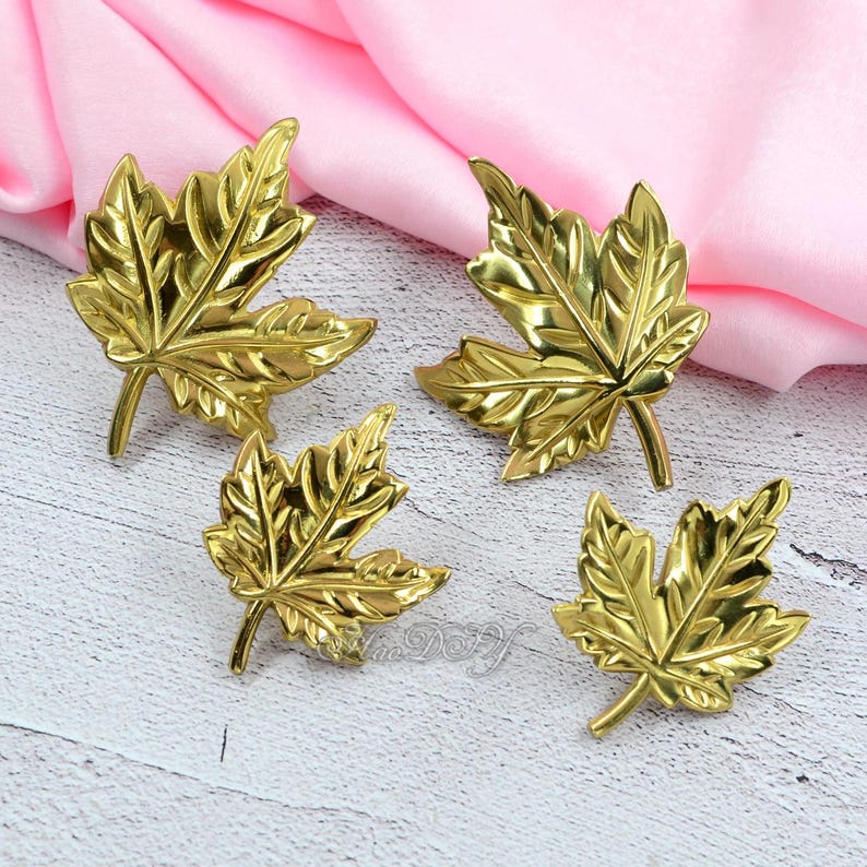 May include: Four gold-colored maple leaf-shaped decorative items. The leaves have detailed veining and a shiny finish. They are arranged on a light-colored wooden surface with a pink fabric in the background. The leaves are likely decorative accents.