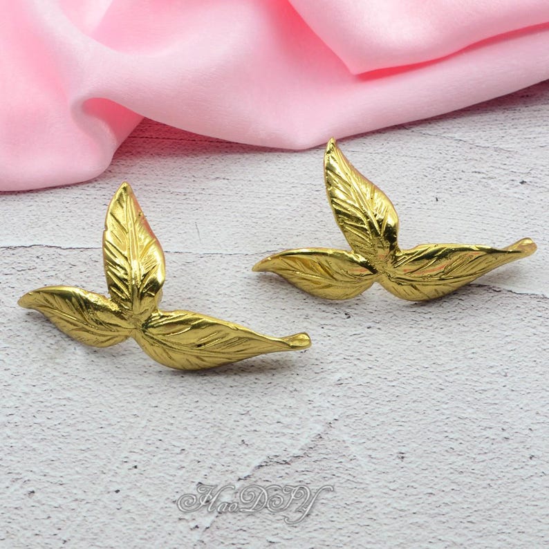 Pu&ograve; includere: Due abbellimenti dorati a forma di foglia. Ognuno ha tre foglie con venature dettagliate. Le foglie sono disposte a ventaglio. Lo sfondo &egrave; una superficie grigio chiaro con un tessuto rosa.