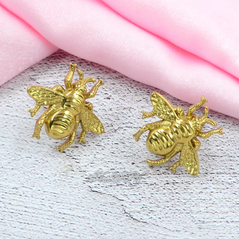 May include: Two gold-coloured bee-shaped ornaments. The bees have detailed wings and legs, and are positioned on a light-coloured surface with a pink fabric in the background. The ornaments are likely jewellery or decorative items.