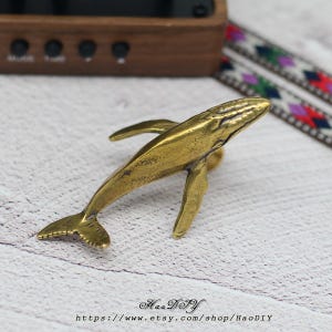 Puede incluir: Una figura de ballena de latón con aletas y cola detalladas. La ballena está en ángulo, mostrando su cuerpo aerodinámico. La figura tiene una estética vintage y es probablemente un artículo decorativo.