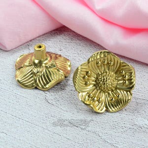 May include: Two gold-colored flower-shaped cabinet knobs. The knobs have detailed petal designs and a central mounting post. The hardware is made of metal and is suitable for furniture or decorative projects.