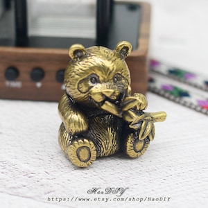 Puede incluir: Pequeña figura de panda dorada, sosteniendo un tallo de bambú en la boca. El panda está sentado, con textura de pelaje detallada y una expresión sonriente. La figura tiene una estética vintage.