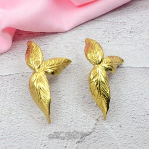 Pu&ograve; includere: Due elementi decorativi dorati a forma di foglia. Ogni foglia presenta venature dettagliate e un design a tre foglie. Le foglie sono esposte su una superficie chiara con un tessuto rosa sullo sfondo.