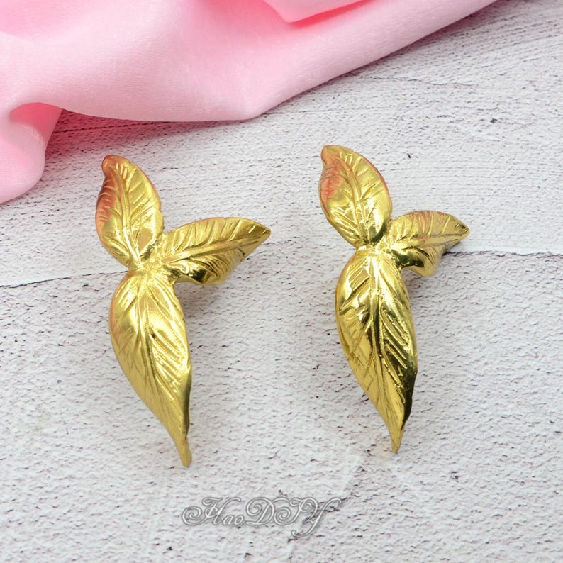 Pu&ograve; includere: Due elementi decorativi dorati a forma di foglia. Ogni foglia presenta venature dettagliate e un design a tre foglie. Le foglie sono esposte su una superficie chiara con un tessuto rosa sullo sfondo.