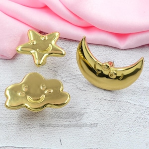 May include: Three gold-colored celestial-themed decorative objects: a smiling star, a smiling cloud, and a crescent moon. The objects are made of a shiny, reflective material and are set against a pink background.