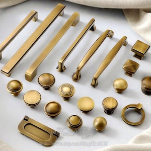 Può includere: Una collezione di ferramenta per mobili in ottone antico, tra cui maniglie, pomelli e un anello di trazione di varie dimensioni. La ferramenta ha una finitura calda e invecchiata, disposta su una superficie neutra. Ideale per il restauro di mobili.