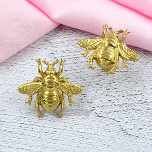 May include: Two gold-coloured bee-shaped decorative objects. The bees have detailed wings, antennae, and body segments. They are displayed on a light-coloured surface with a pink fabric in the background.