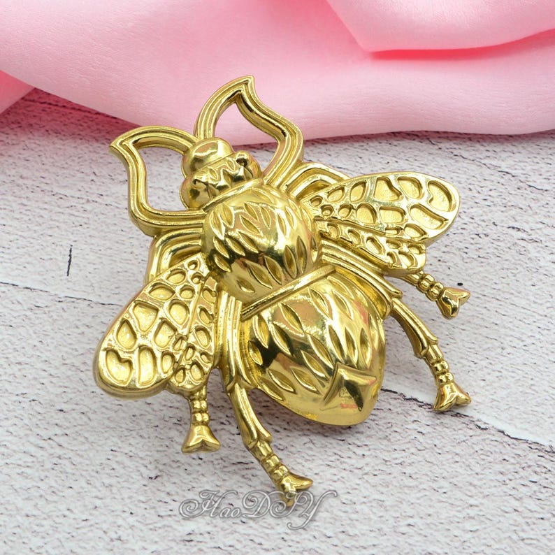 Peut inclure: Une broche dor&eacute;e en forme d'abeille avec des d&eacute;tails complexes sur les ailes, le corps et les pattes. La broche a une finition polie et est pos&eacute;e sur un fond rose et blanc. Le texte "Hao D&F" est en bas.