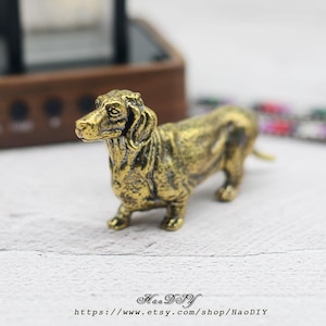 May include: A detailed, gold-toned dachshund ornament. The dog is depicted standing, with a textured finish and lifelike details. The figurine is on a white surface. The Etsy shop name and URL are visible at the bottom of the image.