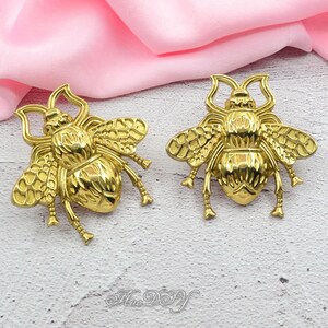 Peut inclure: Deux ornements d&eacute;coratifs en forme d'abeille de couleur or. Les abeilles ont des ailes, des pattes et des antennes d&eacute;taill&eacute;es. Le fond est un tissu rose tendre. Les ornements sont probablement destin&eacute;s &agrave; l'artisanat ou &agrave; la bijouterie.