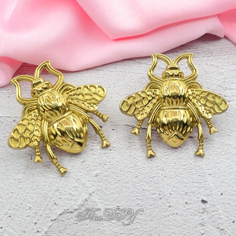 Peut inclure: Deux ornements d&eacute;coratifs en forme d'abeille de couleur or. Les abeilles ont des ailes, des pattes et des antennes d&eacute;taill&eacute;es. Le fond est un tissu rose tendre. Les ornements sont probablement destin&eacute;s &agrave; l'artisanat ou &agrave; la bijouterie.