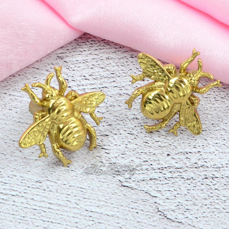 May include: Two gold-coloured bee-shaped decorative objects. The bees have detailed wings, antennae, and body segments. They are positioned on a white and grey surface with a pink fabric in the background.