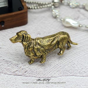 May include: A detailed, golden-colored dachshund figurine. The dog is in a standing position, with its legs visible. The figurine has a textured surface and is likely made of metal. The Etsy shop name and URL are visible at the bottom.