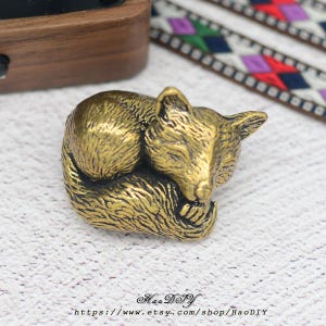Puede incluir: Una detallada figura de zorro dorada en posición fetal durmiendo. El zorro es de metal y tiene detalles intrincados. El nombre y la URL de la tienda Etsy son visibles en la parte inferior de la imagen.