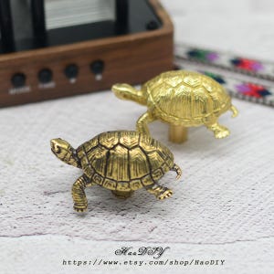 Op de afbeelding: Twee messingkleurige schildpadbeeldjes. De schildpadden hebben gedetailleerde schildpatronen en staan in een loopstand. De beeldjes staan op een wit oppervlak. De Etsy shopnaam en URL zijn zichtbaar onderaan de afbeelding.