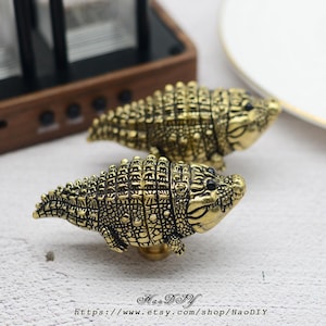 May include: Two brass-colored crocodile-shaped cabinet knobs. The knobs have detailed scales and black eyes. The knobs are on a light-colored surface. The Etsy shop name and URL are printed at the bottom of the image.