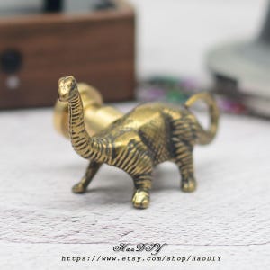 May include: A small, brass-coloured dinosaur figurine with a long neck and tail. The figurine has detailed texture and is posed in a standing position. The Etsy shop name and URL are visible at the bottom of the image.