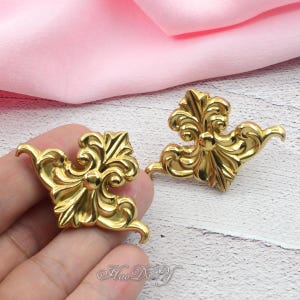 May include: Two ornate, gold-colored decorative furniture corner pieces. Each piece features a symmetrical design with swirling patterns and a central rounded element. The pieces are likely made of metal and are displayed against a pink and white background.