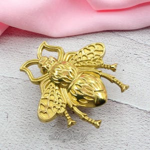 Peut inclure: Un objet d&eacute;coratif dor&eacute; en forme d'abeille. L'abeille pr&eacute;sente des ailes, des pattes et des segments de corps d&eacute;taill&eacute;s. L'objet est probablement en m&eacute;tal et a une finition brillante. L'image est prise en gros plan.