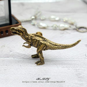 Op de afbeelding: Een gedetailleerd messingkleurig Tyrannosaurus Rex beeldje. De dinosaurus staat met zijn staart uitgestrekt en zijn armen langs zijn zij. Het beeldje heeft een textuur en staat op een wit oppervlak. De tekst "HaoDIY" en een websiteadres staan onderaan.