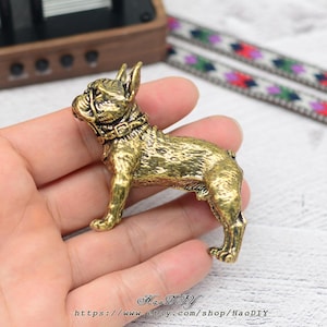May include: A golden-coloured French Bulldog figurine. The dog is shown standing, wearing a collar. The figurine has a textured surface, possibly metal. The Etsy shop name and URL are visible at the bottom of the image.
