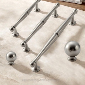 May include: A collection of silver-toned cabinet hardware, including elongated bar pulls and spherical knobs. The pulls have a cylindrical center with rounded ends and are mounted on small, circular bases. The knobs are spherical and also sit on circular bases.