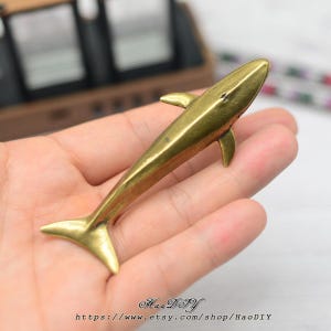 Könnte beinhalten: Eine kleine, messingfarbene Hai-Figur mit Flossen und Schwanz. Der Hai wird in einer Hand gehalten, was sein detailliertes Design zeigt. Der Name und die URL des Etsy-Shops sind unten im Bild aufgedruckt.