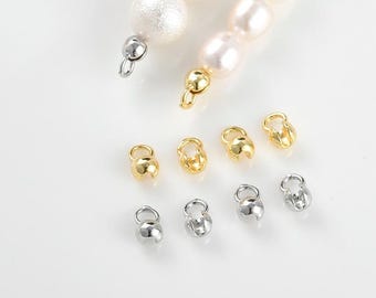 May include: An overhead shot displays a collection of jewelry findings in gold and silver tones. The findings are small, bell-shaped clasps, some attached to a strand of pearls. The pearls are white and round, with a lustrous sheen.