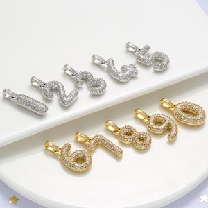 Może przedstawiać: Zestaw dziesięciu złotych i srebrnych zawieszek w kształcie cyfr od 1 do 10. Każda zawieszka jest pokryta małymi, przezroczystymi cyrkoniami.