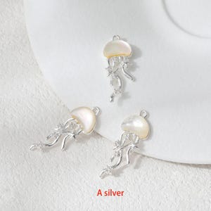 May include: Three silver-colored jellyfish charms with pearlescent tops and dangling tendrils. The charms have a loop at the top for attaching to jewelry. The text "A silver" is at the bottom of the image.